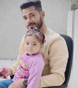 Photo of a man in a beige sweatshirt and holding his young daughter. The girl is wearing a pink sweatshirt and has clips in her hair.