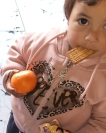 Photo of a young child with brown eyes and hair in a pink hoodie. The child has a cookie in their mouth and a citrus fruit in their hand.